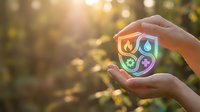 Hand holding glowing shield icon with fire, water, leaf, and recycling symbols representing renewable energy and sustainable environmental protection outdoors.