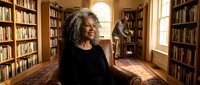 Mature African American woman with curly gray hair smiling warmly in cozy home library with wooden bookshelves and natural lighting for lifestyle content.