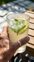 Caucasian male hand holding a refreshing lemonade cocktail with fresh mint leaves and lemon slices, ice cubes, outdoors on wooden deck during summer day.