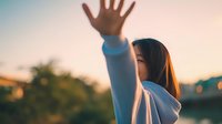 Young Asian woman raising hand against sunset sky, expressing freedom, hope, or farewell in warm golden hour light.