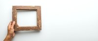 Hand holding empty rustic wooden picture frame against white wall background with copy space for design and text placement.