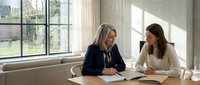 Two professional businesswomen having meeting at modern office table discussing documents and paperwork in bright workspace with large windows.
