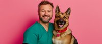 Cheerful veterinarian in green scrubs embracing happy German Shepherd dog with red collar against pink background, showing positive pet care relationship.
