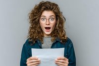 Young woman with curly hair and glasses expressing shock while holding document against gray background, portraying surprise reaction.