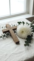 Wooden cross with white chrysanthemum flower and eucalyptus branches on soft fabric near window creating peaceful memorial or religious composition.