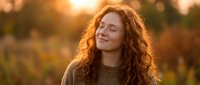 Young redhead woman with curly hair smiling peacefully with eyes closed in warm golden sunlight during autumn season outdoors.