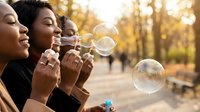 Three diverse women enjoying outdoor bubble blowing activity in warm golden sunlight creating joyful friendship moment in park setting during leisure time.