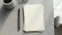 Blank white paper sheets stacked on speckled gray surface with pen and coffee cup for writing, planning, and creative work projects.
