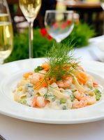 Creamy pasta with shrimp, green peas and fresh dill garnish served on white plate. Wine glasses and natural greenery create elegant restaurant atmosphere.