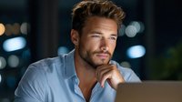 Handsome young Caucasian businessman with beard working late on laptop in modern office with city lights background for corporate and professional content.