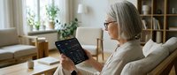 Senior woman with glasses using digital tablet for financial trading and investment analysis while relaxing in comfortable modern living room at home.