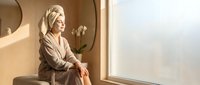 Relaxed woman in bathrobe sitting by window enjoying peaceful morning light in modern bathroom spa setting for wellness and self-care routine.