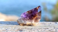 Raw amethyst crystal point with deep purple and brown zones on rough base, illuminated by natural sunlight against blurred outdoor background.