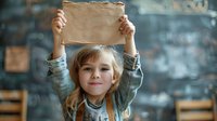 Young girl with blonde hair holding blank cardboard sign overhead, smiling at camera against blurred chalkboard background in classroom setting.