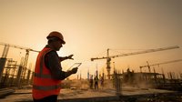Male South Asian construction engineer in hard hat and safety vest using tablet at building site during golden sunset.