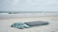 Laptop wrapped in light blue towel lying on sandy beach with ocean waves and cloudy sky in background, representing work-life balance, remote work, vacation, and digital detox concepts.