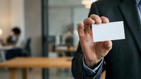 Businessman in dark suit holding blank white business card in modern office environment with blurred colleagues working in background.