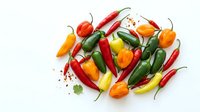 Assorted fresh chili peppers and bell peppers arranged on white background with scattered seeds and herbs, suitable for food photography, cooking blogs, and culinary design projects.