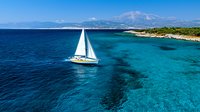 Aerial view of white sailboat cruising through crystal clear turquoise Mediterranean waters near coastal shoreline with mountains in background on sunny day.