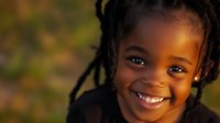 Joyful African American young girl with braided hair smiling brightly at sunset, natural warm lighting creates golden glow on her face, expressing pure happiness.