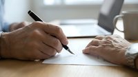 Senior male professional with light skin tone writing or signing document at wooden desk in modern office environment with coffee cup and blurred background.