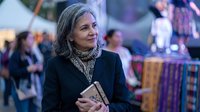 Mature woman with gray hair smiling at outdoor market, holding book, wearing elegant dark coat. Professional lifestyle portrait for business content.