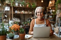 Mature woman in glasses and headphones working on laptop in cozy home garden room with plants. Remote work lifestyle of senior female entrepreneur.