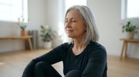 Peaceful mature woman with gray hair meditating with closed eyes in bright modern living room, practicing mindfulness and relaxation at home.