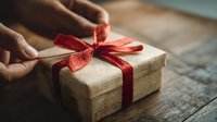 Hands holding rustic gift box wrapped in burlap with red ribbon bow on wooden table. Perfect for holiday, birthday, or anniversary celebration.