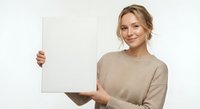 Young blonde Caucasian woman in beige sweater holding blank white sign with both hands, smiling at camera against light background for mockup designs.