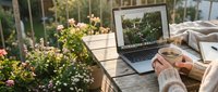 Young woman working on laptop outdoors on sunny balcony with coffee cup, flowers blooming around wooden table in warm morning light.