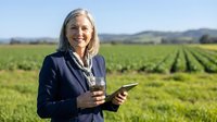 Confident senior Caucasian woman agronomist holding tablet and soil sample in green farm field, smiling outdoors in blue blazer.