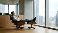 Young Asian businessman relaxing in leather chair with feet up in modern office overlooking city skyline during break time.