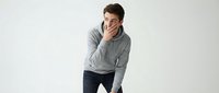 Young man with dark hair and light skin tone wearing casual gray sweatshirt and dark jeans, posing with hand to face in thoughtful gesture against white background for portrait and lifestyle use.