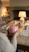 Woman holding pink ceramic piggy bank at rustic wooden dining table in cozy home interior, warm lamp light, savings and financial planning concept.