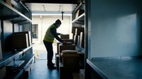 Warehouse worker in safety vest organizing packages on metal shelving in modern distribution center for logistics and shipping operations.