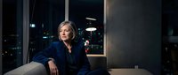 Professional middle-aged Caucasian businesswoman in navy suit sitting confidently in modern office at night with city lights background for corporate content.
