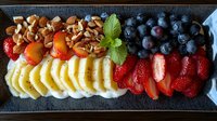 Fresh fruit platter with sliced bananas, strawberries, blueberries, kumquats and roasted nuts arranged in colorful rows on dark plate garnished with mint leaf.