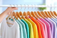 Colorful rainbow t-shirts hanging on wooden hangers in closet, arranged by color from white through pastels to bright shades. Organized wardrobe storage solution.