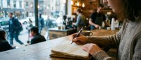 Woman in sweater writing in notebook at cafe table with urban street view through window, suitable for lifestyle, business, and creative work concepts.