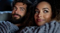 Young diverse couple relaxing under cozy knit blanket at home, sharing intimate moment while watching movie in dark room with soft lighting.