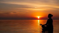 Woman meditating in lotus position by calm water during golden sunset with warm orange sky reflecting on surface creating peaceful wellness moment.