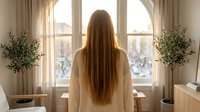 Young woman with long brown hair in white robe standing by large window overlooking city street at sunrise, peaceful morning routine in modern apartment.