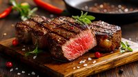 Grilled medium-rare steak sliced on wooden board with sea salt, fresh herbs, and red chili peppers, showcasing juicy meat interior for fine dining and culinary content.
