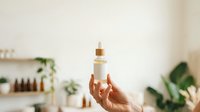 Cropped hand holding white cosmetic serum bottle with gold dropper cap against blurred spa background with plants and skincare products for beauty and wellness marketing.