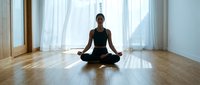 Young woman of South Asian descent in black athletic wear practicing meditation in lotus position on wooden floor with natural light from large windows and sheer curtains.