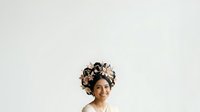 Young woman with medium skin tone wearing an elaborate floral headwrap featuring black and cream-colored flowers, isolated on white background for design and editorial use.
