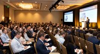 Business conference with speaker presenting to large audience in modern hotel ballroom with professional lighting and projection screens.