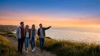 Group of four young adults standing on coastal cliff at sunset, enjoying scenic ocean view and golden sky. Perfect for travel, lifestyle, and tourism marketing materials.