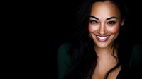 Young mixed race woman with long dark hair and bright genuine smile wearing green top against black background, dramatic studio lighting creates professional portrait.